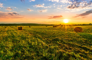 Scenic view at picturesque burning sunset in a green shiny field with hay stacks, bright cloudy sky , trees and golden sun rays, summer valley landscape © Yaroslav