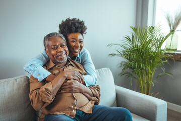Senior man and his middle aged daughter smiling at each other embracing, close up. Portrait of a daughter holding her elderly father, sitting on a bed by a window in her father's room.