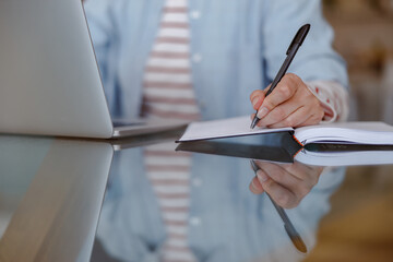 Naklejka premium Close up of female hand taking notes in notebook while woman sitting at glass table with modern laptop