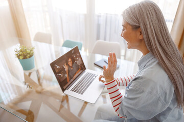 Female person sitting at the table with laptop and doing hello gesture while talking with friend through video call