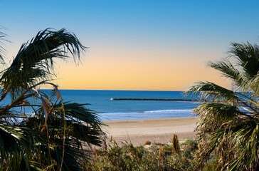 Empty beach on the sea with palm trees. Stone pier on coastline from to side Playa Puebla de Farnals. Coast at sea with waves on sunset sky background. Sea and palms at coastline in Spain. .
