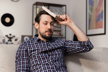 A man tries to memorize new English words. A bearded man sits on the couch and hits his head with a dictionary, hoping that he will finally remember the definition.