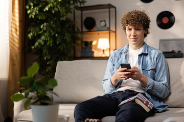 A boy uses a smartphone while teaching English at home. A young man wearing a denim shirt sits on a couch in the living room and reads messages on a smartphone screen.