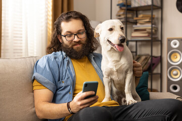 A bearded man browses the internet on a smartphone in the company of his dog. A contented middle-aged boy uses his phone while sitting on the living room couch.