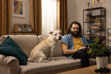 A bearded man spends his free time with his dog at home. The boy is holding a phone in his hands and looking towards the dog. Leisure time with his canine friend.