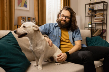 A bearded boy spends his free time with his dog while sitting on the couch in the living room.