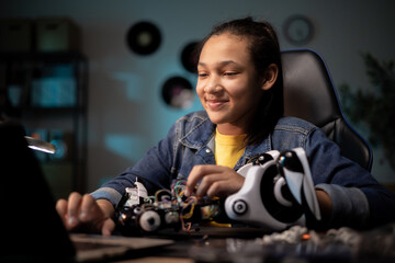 Cheerful girl sits at desk in room in the evening, scrolling on laptop, preparing the programming that controls the robot, passionate about robotics, electronics, computer science