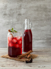 A transparent glass with red tea, cinnamon and ice on a wooden table. A bottle of drink in the background. Cinnamon tubes and dried hibiscus flowers on a canvas napkin