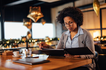 African-American woman with glasses, looking at the menu while sitting.