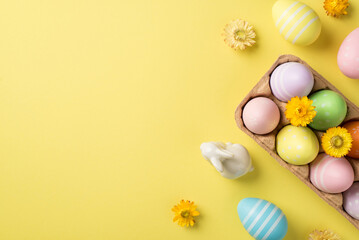 Top view of a half carton with multicolored Easter eggs and flowers white figure of rabbit and three pastel eggs and bright flowers on the isolated yellow empty background