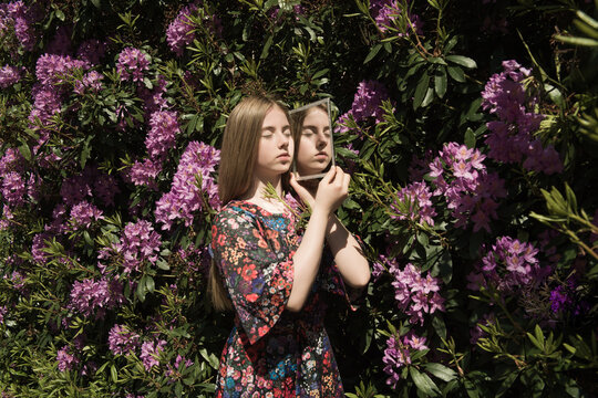 girl in floral dress holding mirror in the garden near pink and purple rhododendron bush