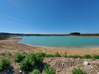 Das Blau des Himmels spiegelt sich im Wasser des Sees. Der See ist von einem Sandstrand umgeben. Grüne Unkraut-Büsche aus dem Sand. Die Bäume sind wie ein Wald gruppiert. Raum für Ruhe. Erholung.