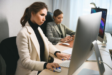Fototapeta premium Two young adult women work in an office or coworking space on computers. Two casual businesswomen at work in a modern office space