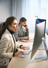 Two young adult women work in an office or coworking space on computers. Two casual businesswomen at work in a modern office space