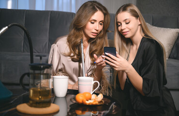 Girls party at home. Group of two young women in backlight smoking shisha in cafe or bar, making smoke clouds, having fun, smiling. Relax concept. Friendship