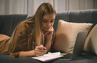 A teenager girl is engaged in homeschooling using a laptop, making notes in a notebook. Study on the couch, distance learning and work