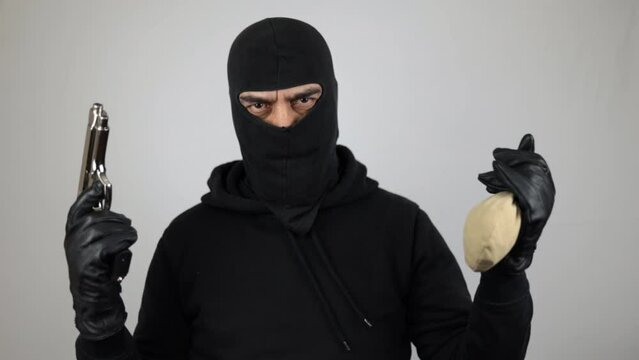 Man with black balaclava holding the pistol and a bag full of jewelry, over white background. Crime concept