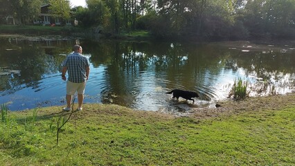 Obraz na płótnie Canvas Mensch und Hund am Ufer des Sees. Der Hund läuft auf dem Wasser. Es ist ein sonniger Tag am Fluss. Der schwarze Labrador liebt es zu schwimmen. Das Gras am Ufer ist grün und dicht. Das Wald-Reflexions