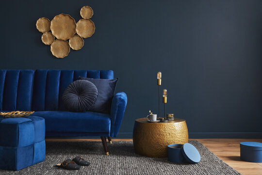 Stylish modern living room interior design with glamour blue velvet sofa, pouf, golden metal side table and modern home accessories. Dark blue wall. Template. Copy space. © FollowTheFlow