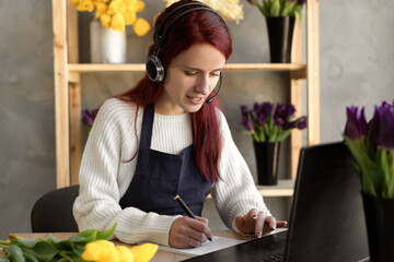 Young female florist in blue apron business owner using headset and computer while talking to...