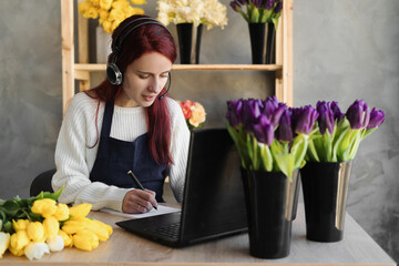 Young woman florist in blue apron business owner using headset and computer while talking with customers online takes order online, write down with pen in notebook.