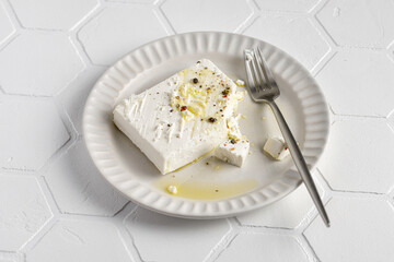 Homemade marinated feta cheese on ceramic white plate with olive oil and pepper. Tile background