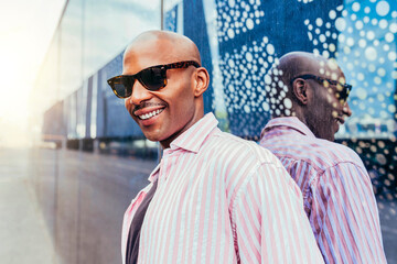 Portrait of cheerful young mixed race stylish homosexual bald man wears sunglasses, leaning against...