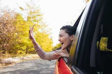 Happy smiling young woman looking out of car window on the go and waving - obrazy, fototapety, plakaty