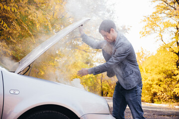 A man looks under the open hood of a car. The car broke down on the road. The engine is smoking
