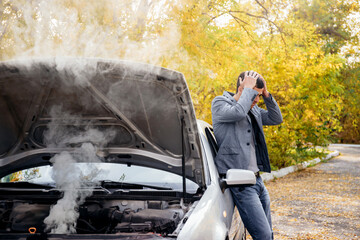 A man looks under the open hood of a car. The car broke down on the road. The engine is smoking