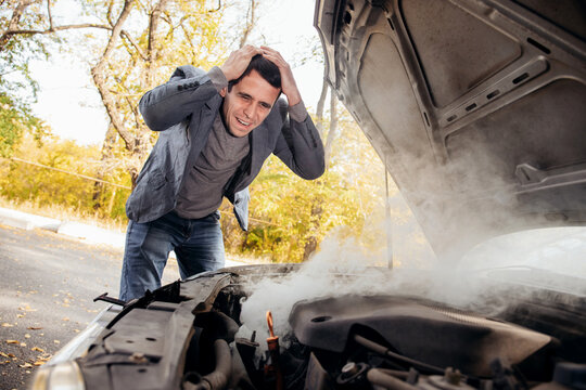 A Man Looks Under The Open Hood Of A Car. The Car Broke Down On The Road. The Engine Is Smoking