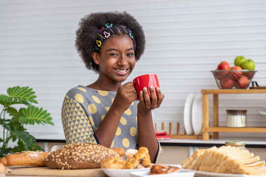 Young beautiful African black girl woman sitting in kitchen with breakfast, bread and egg, holding coffee mug.