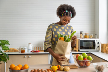 Young beautiful African black girl woman in kitchen holding paper grocery shopping bag full of fresh healthy food, fruit and vegetable.