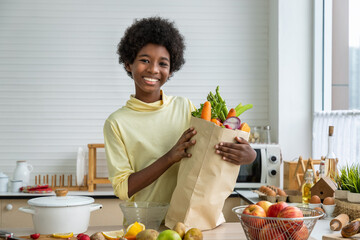 Young cute African black boy in kitchen holding paper grocery shopping bag full of fresh healthy...