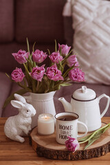 Still-life. A vase with flowers, tulips, a teapot, a bunny, a candle and a cup of tea on the coffee table in the living room home interior. Cozy spring concept.