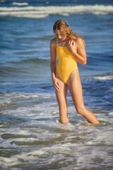 A young woman in a yellow swimsuit wades slowly in the water at the beach