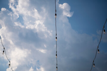 Light bulbs on string wire against beautiful blue cloudy sky background. Long garland of lights. A wedding, event or festival banquet decoration.