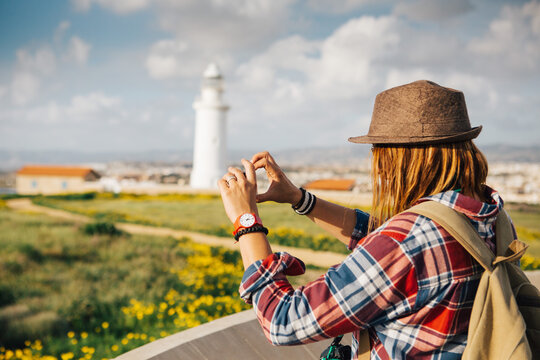 A young woman traveler in a hat and with a backpack in front of a strictly archaeological site with a lighthouse. She holds the phone in her hand and takes pictures. Sunny day, spring, Cyprus.