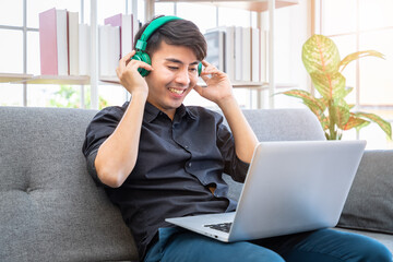 Asian businessman freelance sitting on sofa in living room using compute laptop with headphone. Concept for working at home.