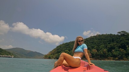 Pretty young blonde woman in stylish swimsuit and sunglasses sits on pink canoe sailing along sea...
