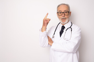 Senior grey-haired doctor man standing over isolated white background surprised face pointing finger up looking at the camera. Positive person.