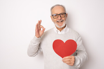 Happy senior man showing red paper heart and with okay sign isolated over white background. Health...