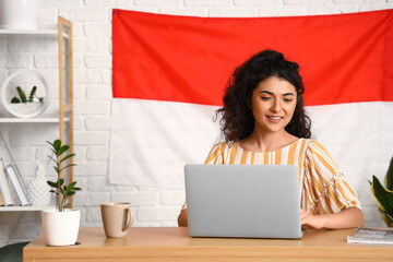 Asian woman sitting at table with modern laptop in room with big Indonesian flag