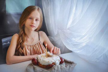A girl with long blond hair looks into the frame, a birthday cake, a cupcake stands nearby....