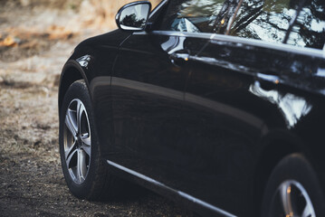 A black vehicle stands on a forest road. The vehicle is photographed from the left rear side. The wheels get dirty lightly from the mud.
