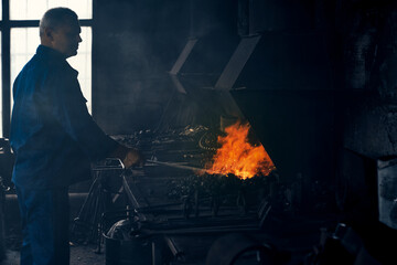 Side view of worker in dark blue suit standing near special heating equipments and working with metal in smithy. Concept of process creating modern fence.  - Powered by Adobe