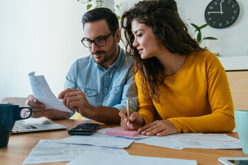 Smiling Loving Couple Paying Bills at Home. 
Smiling boyfriend and girlfriend sitting at desk together and using laptop computer and calculator while holding bills and calculating family budget.