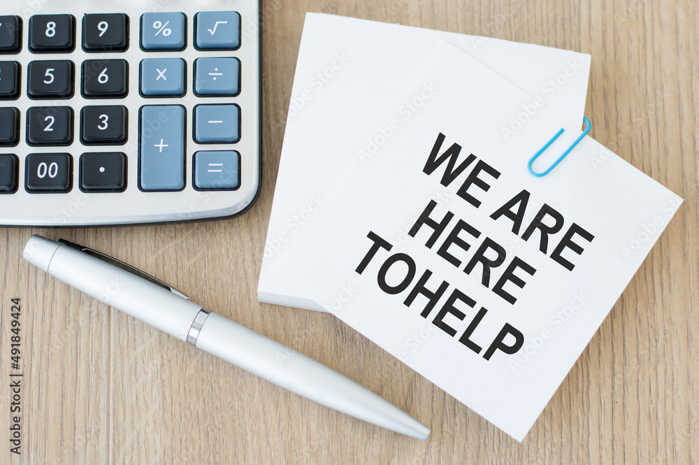 Poster text we are here to help on a card on a paperclip on a wooden table next to a pen and calculator