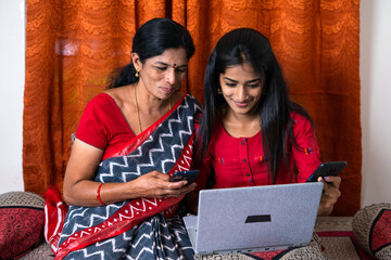 Mother and teenager daughter using mobile phone while watching taptop at home - concept of technological lifestyle, social media distaraction and using multiple gadgets