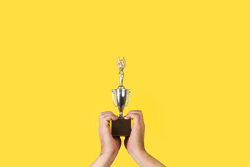 Manos de hombre sosteniendo y levantado un trofeo sobre un fondo amarillo liso y aislado. Vista de frente. Copy space
